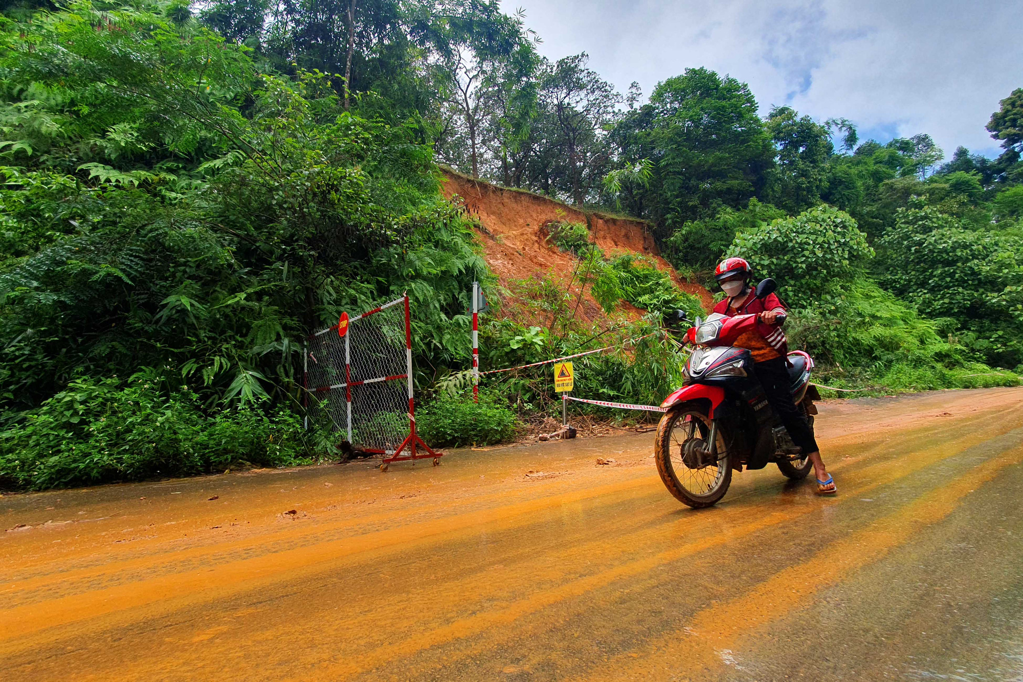 'Thót tim' qua điểm sạt lở trên tuyến đường nối Bình Phước với Lâm Đồng - Ảnh 5. Hàng chục khối đất đá từ đỉnh đồi ta luy dương đổ tràn xuống lấp mương thoát nước ven đường và lòng đường. Mưa tạo thành lớp bùn đỏ, trơn trượt, người đi xe hai bánh qua lại rất khó khăn. Trong ảnh: một phụ nữ phải rà chân để đi qua đoạn đường sạt lở trơn trượt sau cơn mưa