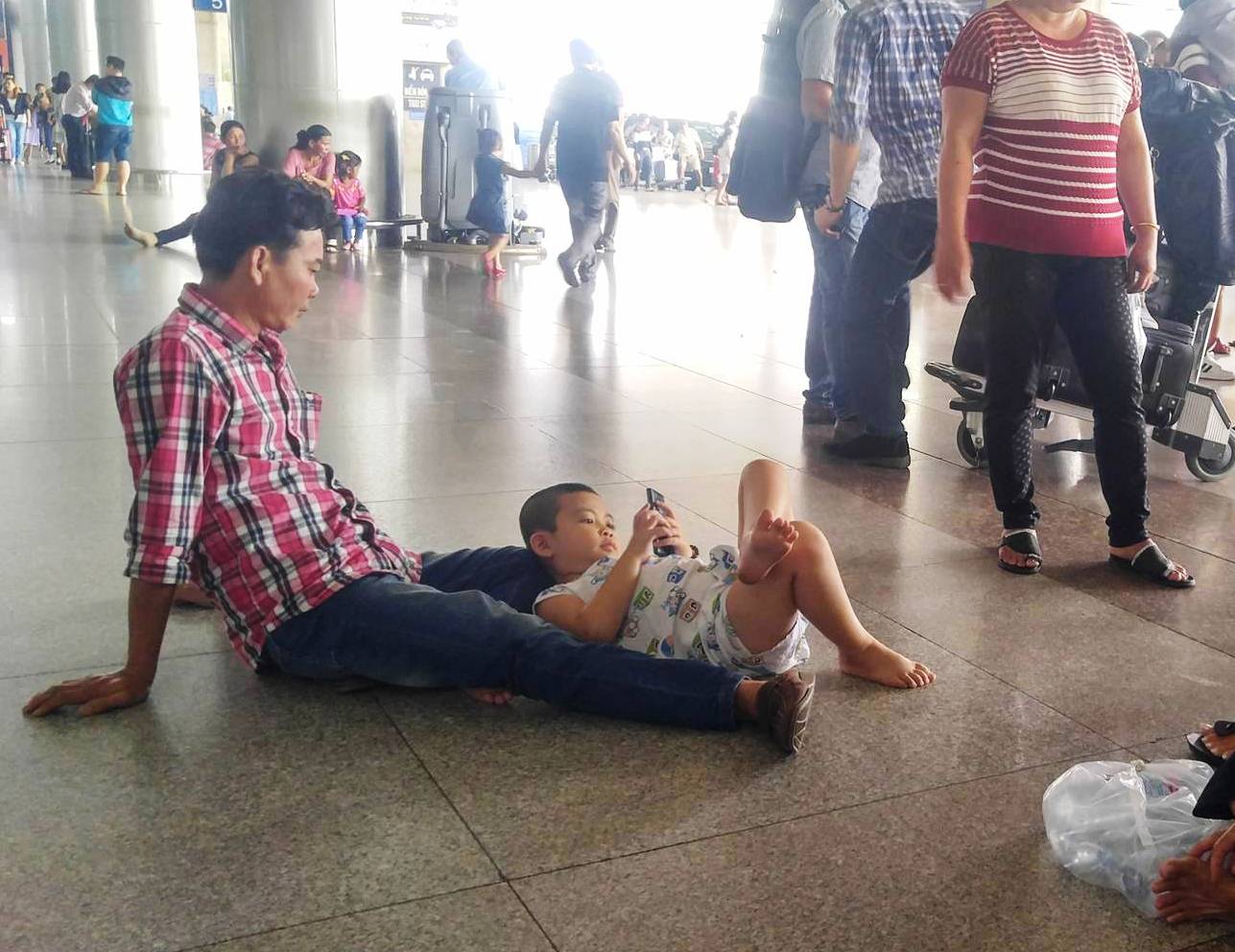 Hai cha con ngồi bệt xuống nền nhà ga bấm điện thoại trong lúc chờ đợi
