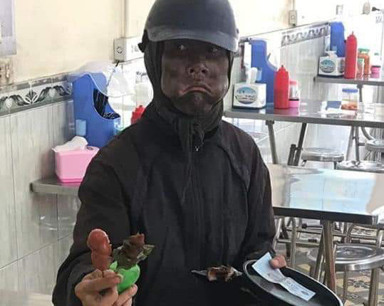 Người đàn ông bôi đen mặt, phùng má, tay cầm đầu gà đi lang thang xin tiền