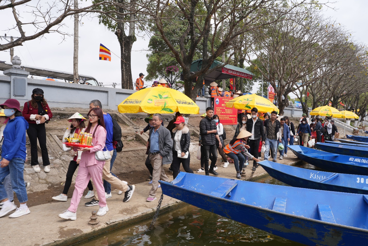 A group of people walking by a canal with boats and umbrellas  Description automatically generated