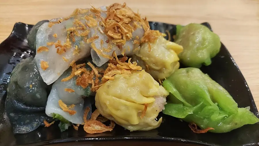 Những ng&agrave;y mưa, khu Chợ Lớn lu&ocirc;n c&oacute; một kiểu hấp dẫn rất ri&ecirc;ng đến từ những xửng h&aacute; cảo, x&iacute;u mại bốc kh&oacute;i nghi ng&uacute;t b&ecirc;n lề đường. Chỉ cần đứng gần l&agrave; đ&atilde; nghe m&ugrave;i bột hấp n&oacute;ng quyện c&ugrave;ng nh&acirc;n t&ocirc;m thịt thơm lừng. Từng vi&ecirc;n h&aacute; cảo trắng trong, lớp vỏ mềm dai &ocirc;m lấy phần nh&acirc;n đậm đ&agrave;; x&iacute;u mại th&igrave; b&eacute;o thơm, ăn k&egrave;m nước tương pha sa tế cay nhẹ c&agrave;ng bắt vị. Đ&acirc;y l&agrave; m&oacute;n kh&ocirc;ng qu&aacute; no nhưng lại rất hợp để vừa tr&uacute; mưa vừa nhẩn nha thưởng thức. Cảm gi&aacute;c mở nắp xửng, l&agrave;n kh&oacute;i n&oacute;ng phả l&ecirc;n mặt giữa tiết trời se lạnh khiến m&oacute;n ăn d&acirc;n d&atilde; n&agrave;y bỗng trở n&ecirc;n cuốn h&uacute;t lạ thường.