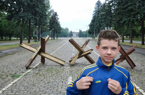 Mặc kệ chiến tranh, người dân Ukraine hòa vào không khí Euro 2024- Ảnh 1. Một cậu bé người Ukraine khoe bộ đồ thể thao mới của đội tuyển quốc gia - món quà em được mẹ tặng trước mùa giải Euro 2024. Phía sau là nhím chống tăng được thiết lập để cản đường xe tăng tại các con đường chính xung quanh nhà ga trung tâm thành phố Odessa và khu vực Kulykove Pole - Ảnh: RICHARD MORGAN