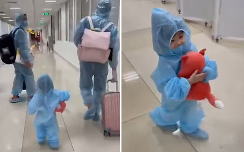 Em bé mặc đồ bảo hộ, ôm gấu bông vui vẻ cùng bố mẹ đi cách ly